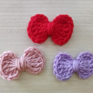 May include: Three crocheted bows in different colors: red, pink, and purple. The bows are made with a textured yarn and have a simple, classic design.