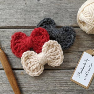 May include: Three crocheted bows in red, dark gray, and cream colors. A wooden crochet hook, a ball of cream yarn, and a rectangular paper tag with the text "Handmade by eandlcreation" are arranged alongside the bows on a wooden surface.