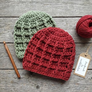 May include: Two handmade crochet beanies in red and green, with a textured waffle stitch pattern. A wooden crochet hook and a ball of red yarn are also visible. The beanies are displayed on a wooden surface.