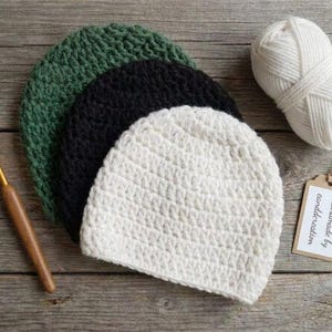 May include: Three crocheted beanies in forest green, black, and cream colors. A wooden crochet hook, a ball of cream yarn, and a rectangular paper tag with the text "Handmade by eanddcreation" are arranged alongside the hats.