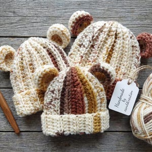 May include: A collection of handmade crochet bear hats in cream, brown, and tan, with bear ears. The hats are displayed on a wooden surface, alongside a ball of yarn and a crochet hook. A tag reads "Handmade by canddcreation."