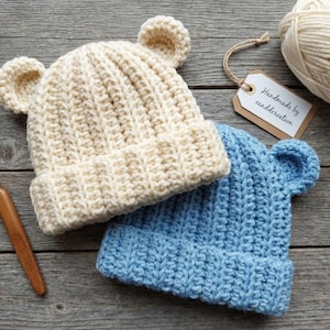 Puede incluir: Dos gorros de oso de ganchillo hechos a mano en crema y azul claro, con detalles de orejas a juego. Un ganchillo de madera, una madeja de hilo crema y una etiqueta que dice "Handmade by canddcreation" también son visibles. Sobre una superficie de madera envejecida.