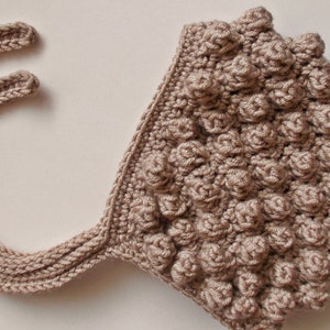 Puede incluir: Un gorro de bebé de ganchillo beige con un diseño texturizado y abultado. El gorro tiene dos lazos largos.