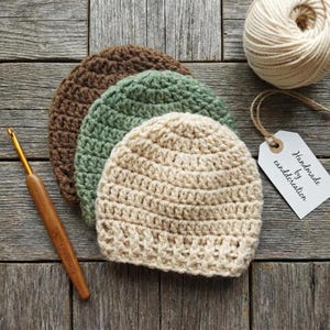 May include: Three crocheted beanies in brown, sage green, and cream, stacked together. A wooden crochet hook lies to the left, and a ball of cream yarn with a white tag reading "Handmade by eandcreation" is positioned to the right.