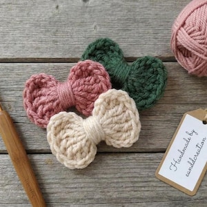 May include: Three crocheted bows in dusty pink, dark green, and cream yarn. A wooden crochet hook, a ball of dusty pink yarn, and a rectangular paper tag with the text Handmade by canddcreation are arranged alongside the yarn bows.