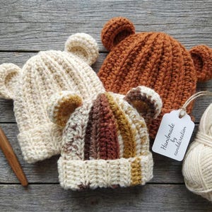 May include: Three handmade crochet bear hats in cream, brown, and a multi-colour pattern. A ball of cream yarn, a crochet hook, and a tag that says "Handmade by canddcreation" are also in the image. The hats have bear ears.