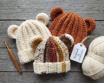May include: Three handmade crochet bear hats in cream, brown, and a multi-colour pattern. A ball of cream yarn, a crochet hook, and a tag that says "Handmade by canddcreation" are also in the image. The hats have bear ears.