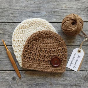 May include: Two crocheted beanies, one cream and one brown, featuring a dark brown wooden button on the brown hat. A wooden crochet hook lies to the left, and a ball of brown yarn with a white tag labeled "Handmade by candcreation" is included.