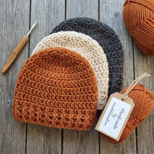 Può includere: Berretti all'uncinetto fatti a mano in ruggine, crema e grigio antracite. Sono visibili anche un uncinetto e due gomitoli di lana. Un'etichetta recita "Handmade by candicreation."