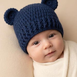 May include: A dark navy blue crochet hat featuring two small, rounded bear-like ears on top. The hat is constructed with a textured stitch pattern and has a ribbed brim that fits snugly around the head, providing a simple and functional design.