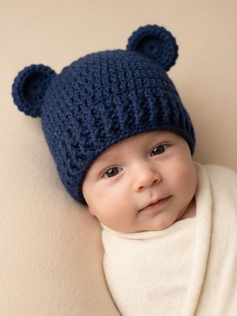 May include: A dark navy blue crochet hat featuring two small, rounded bear-like ears on top. The hat is constructed with a textured stitch pattern and has a ribbed brim that fits snugly around the head, providing a simple and functional design.