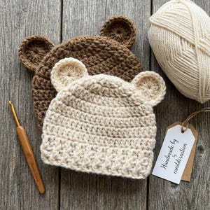 Pode incluir: Dois chapéus de urso de crochê em creme e castanho, com orelhas presas, são exibidos em uma superfície de madeira. Uma bola de lã creme, uma agulha de crochê e uma etiqueta com "Handmade by canddcreation" também estão presentes.