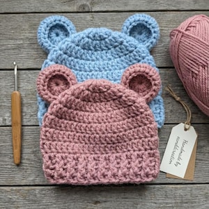 May include: Two crocheted beanies with rounded ears, one light blue and one dusty pink, stacked on top of each other. A wooden crochet hook, a ball of dusty pink yarn, and a white rectangular tag with the text Handmade by eanddcreation are included.