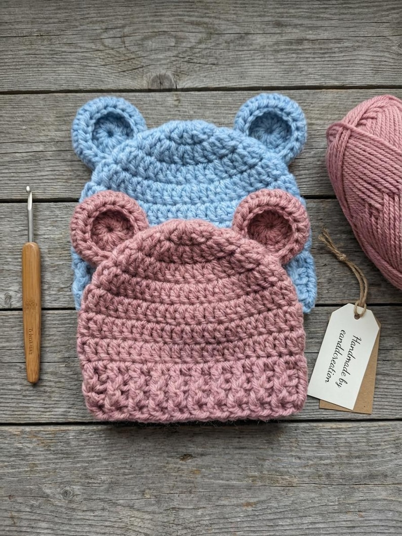 May include: Two crocheted beanies with rounded ears, one light blue and one dusty pink, stacked on top of each other. A wooden crochet hook, a ball of dusty pink yarn, and a white rectangular tag with the text Handmade by eanddcreation are included.