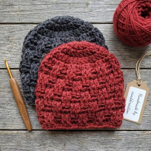 May include: Two handmade crochet beanies in gray and red-orange, with a ball of red yarn and a crochet hook. The beanies have a textured stitch pattern. A tag reads "Handmade by eanddcreation".