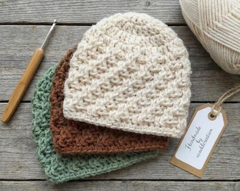 May include: Three crocheted beanies in cream, brown, and sage green stacked together. A wooden crochet hook lies to the left, a ball of cream yarn sits at the top right, and a rectangular tag with the text Handmade by is attached by twine.