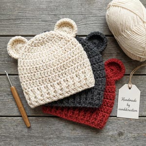 May include: Three crocheted hats with bear ears in cream, charcoal gray, and red. A wooden crochet hook lies to the left, and a ball of cream yarn sits at the top right. A white tag attached to the red hat reads Handmade by eanddcreation.