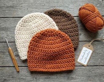 May include: Three crocheted beanies in cream, dark brown, and burnt orange, accompanied by a matching ball of burnt orange yarn, a wooden crochet hook, and a rectangular paper tag with the printed text Handmade by eanddcreation.