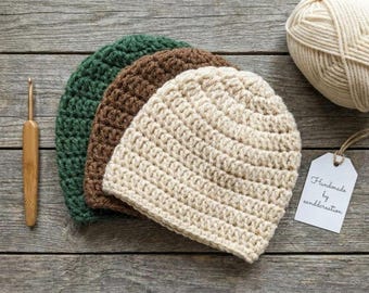 May include: Three crocheted beanies in dark green, brown, and cream colors. A bamboo crochet hook, a ball of cream yarn, and a white tag with the text Handmade by canddcreation are arranged alongside the hats.