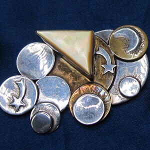 May include: A celestial-themed brooch featuring a triangular mother-of-pearl center, surrounded by silver, gold, and bronze metal elements. The design includes circular shapes, shooting stars, and crescent moons, creating a unique, artistic accessory.