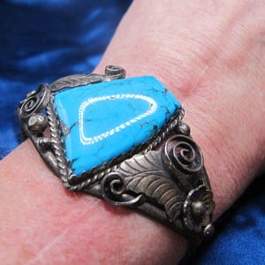 May include: A silver cuff bracelet featuring a large, triangular, bright blue turquoise stone with dark veining. The metal band is decorated with intricate silver leaf motifs, swirling scrollwork, and small spherical accents along the edges.