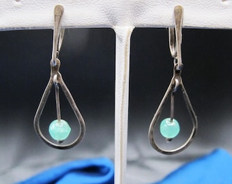 May include: A pair of silver-toned drop earrings. Each earring features a teardrop-shaped frame with a small, round, light green bead suspended in the center. The earrings are displayed against a neutral background.