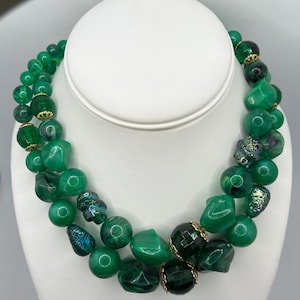 May include: A multi-strand necklace featuring a variety of green beads in different shapes, sizes, and textures. The design includes smooth round beads, faceted dark green beads, and iridescent textured beads, accented with small gold-tone metal caps.