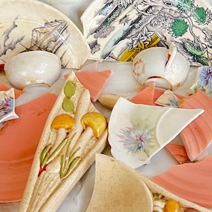 May include: A collection of broken ceramic dishware and teacups. Fragments of plates and cups in shades of peach, white, and beige are scattered across a white surface. Some pieces feature floral and landscape designs.