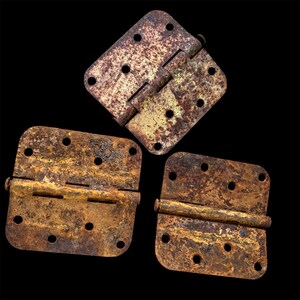 May include: Three rectangular metal door hinges with rounded corners and multiple screw holes. Each hinge displays a heavy, textured brown and orange rust patina covering the original metallic surface, showing signs of age and oxidation.