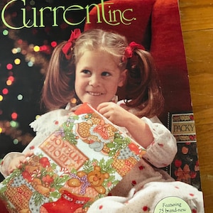 May include: A young girl with pigtails and red bows holds a Christmas-themed gift. The gift is wrapped in colorful paper with teddy bears, stockings, and the words "Joyeux Noel". The background includes the text "Current, Inc."