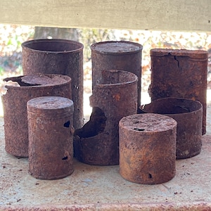 May include: A collection of rusted metal cans of varying heights and diameters. The cans are cylindrical, with visible signs of corrosion and decay. Some cans have holes or are partially broken, showcasing their age and exposure to the elements.