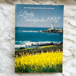 May include: A vintage book titled "Daily Guideposts, 1992" with a coastal scene on the cover. The cover features a lighthouse on a rocky shore, blue ocean, and yellow wildflowers. The text "Spirit-Lifting Thoughts for Every Day of the Year" is also visible.