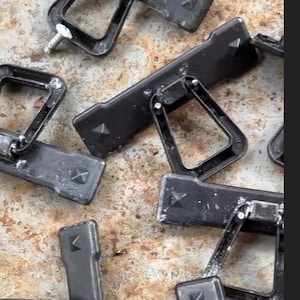May include: A collection of black plastic clips with a triangular design and metal hardware. The clips are scattered on a textured, mottled surface. Some clips have a rectangular base with a raised diamond shape.