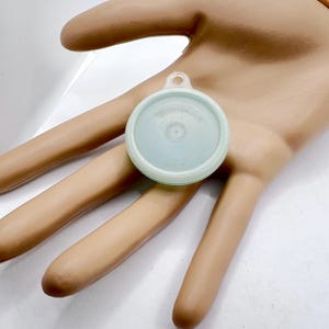 May include: A small, round, pale mint green plastic lid featuring a circular center indentation and a small tab with a hole at the top edge. The lid has a smooth, slightly translucent finish and a raised outer rim, designed for a container.