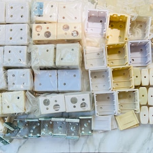 May include: A collection of white and off-white plastic blind mounting brackets, housing components, and small metal screws. The assortment includes various square and rectangular plastic parts, some wrapped in clear plastic, alongside metal hardware.