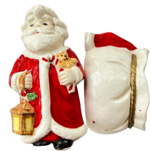 May include: A vintage Christmas decoration featuring a ceramic Santa Claus figure in a red coat with white trim, holding a lantern and a teddy bear. Beside him is a white sack with a red top and gold rope detail.