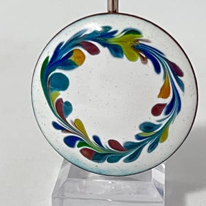 May include: A round, white enamel pendant with a colorful floral design. The design features blue, green, yellow, and red leaf-like shapes arranged in a circular pattern. The pendant has a dark brown border and a silver metal loop.