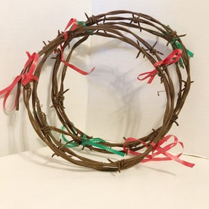 May include: A circular arrangement of rusty barbed wire, adorned with red and green ribbons. The barbed wire is arranged in a layered, wreath-like design, with the ribbons tied in bows at intervals around the circumference.