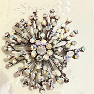 May include: A circular brooch featuring a dark metallic starburst design with numerous radiating arms tipped with iridescent, multi-colored rhinestones. A larger clear rhinestone sits at the center. The brand name Studio 33 is printed above.