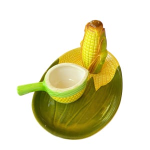May include: A ceramic corn-shaped salt and pepper shaker set with a matching serving dish. The set features a yellow corn cob salt shaker, a small white bowl with a green handle, and a green leaf-shaped dish.