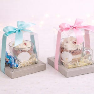 Può includere: Due coppette da dessert con panna montata e torta, presentate in scatole trasparenti con nastri pastello. Ogni coppetta poggia su una base grigia con paglia decorativa e una piccola caramella. Un nastro blu, l'altro rosa.