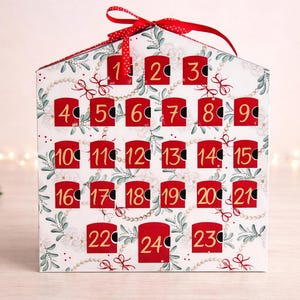 May include: A decorative Christmas advent calendar shaped like a house. The white surface is adorned with red doors numbered 1-24, with a festive pattern of greenery and pearls. A red ribbon is tied at the top.