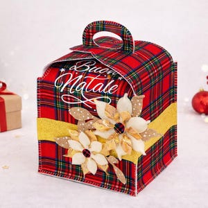 Può includere: Una scatola regalo a quadri rossi con manico, decorata con fiori di stoffa e un nastro dorato. La scatola ha le parole "Buon Natale" in corsivo bianco. Scatola regalo a tema natalizio.