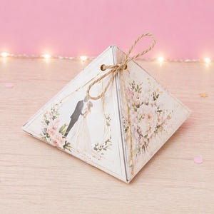 May include: A pyramid-shaped wedding favor box with a floral design and a silhouette of a bride and groom. The box is tied with twine and sits on a wooden surface. Pink confetti and fairy lights are in the background.