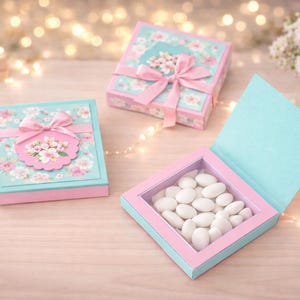 Puede incluir: Tres cajas de regalo decorativas en tonos rosa y azul, adornadas con motivos florales y lazos rosas. Una caja está abierta, revelando caramelos blancos. Las cajas están dispuestas sobre una superficie clara con iluminación suave.