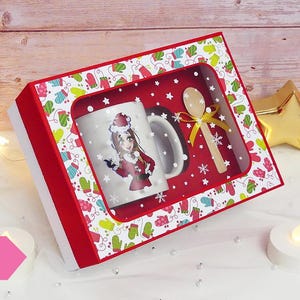 Puede incluir: Una caja de regalo roja y blanca con una ventana transparente que muestra una taza blanca con una imagen de dibujos animados de una persona con un gorro de Papá Noel y un atuendo rojo y blanco. La taza está junto a una cuchara de madera con una cinta dorada. La caja está decorada con un patrón de guantes y copos de nieve.