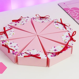 Könnte beinhalten: Acht rosafarbene und weiße Schachteln mit Blumenmuster in Form von Tortenstücken mit roten Schleifen. Die Schachteln sind in einem Kreis auf einer weißen Oberfläche angeordnet. Das Wort "ESEMPIO" ist in schwarzen Buchstaben auf rosa Hintergrund in der linken unteren Ecke des Bildes geschrieben.