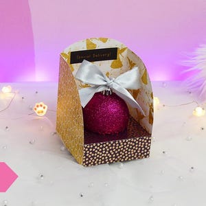 Op de afbeelding: Een roze kerstbal met een zilveren strik wordt getoond in een gouden en bruine kartonnen doos met de woorden "Special Delivery" erop gedrukt. De doos staat op een wit oppervlak met witte lampjes en een wit, pluche oppervlak op de achtergrond.