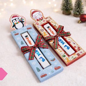 Può includere: Due confezioni regalo festive con barrette di cioccolato Kinder, ciascuna adornata con un nastro a quadri. Una scatola è azzurra con illustrazioni di auto e alberi, l'altra è rossa con un motivo. Topper a forma di pinguino e pupazzo di neve.
