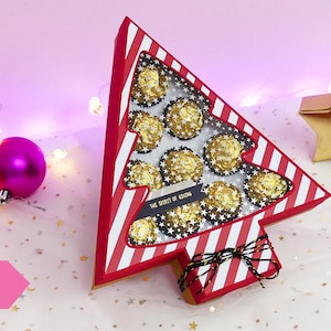 以下が含まれることがあります： 金色のホイルで包まれたチョコレートが、透明な窓から見える、赤と白のストライプのクリスマスツリー型の段ボール箱。黒いリボンと「The Spirit of Giving」の文字が書かれています。